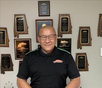 Photo of man in glasses in black collared shirt with SERVPRO logo on the chest in front of a wall with awards hanging